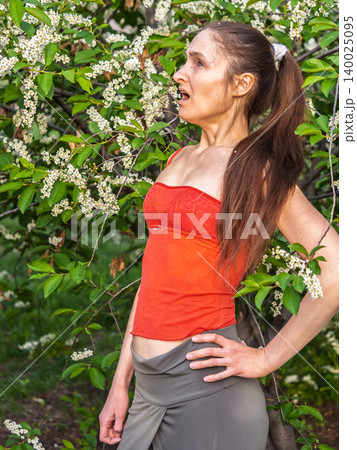 Young woman sneezes in the park against the background of a flowering tree. Allergy to pollen concept. 140025095