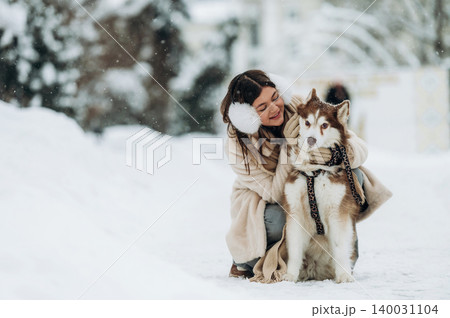Woman with long dark hair wearing earmuffs is hugging a fluffy Siberian Husky dog on a snowy path surrounded by trees during winter season Woman with long dark hair wearing earmuffs is hugging a fluffy Siberian Husky dog on a snowy path surrounded by trees during winter season 140031104