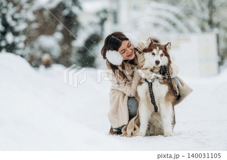 Young woman with brown hair wearing earmuffs is kneeling beside a large Siberian Husky dog in a snowy outdoor setting during winter Young woman with brown hair wearing earmuffs is kneeling beside a large Siberian Husky dog in a snowy outdoor setting during winter 140031105