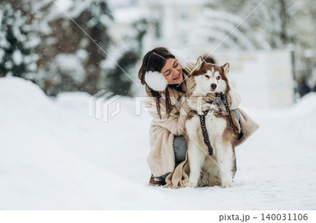 Young girl with long dark hair wearing a winter coat is hugging a fluffy Siberian Husky dog on a snowy path surrounded by trees during a snowfall Young girl with long dark hair wearing a winter coat is hugging a fluffy Siberian Husky dog on a snowy path surrounded by trees during a snowfall 140031106