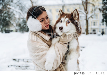 Young woman with long dark hair wearing earmuffs is holding a Siberian Husky dog in a snowy outdoor setting with trees and buildings in the background during winter 140031114