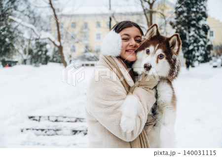 Young woman with long dark hair wearing winter attire is joyfully holding a fluffy Siberian Husky dog in a snowy outdoor setting with trees and buildings in the background Young woman with long dark hair wearing winter attire is joyfully holding a fluffy Siberian Husky dog in a snowy outdoor setting with trees and buildings in the background 140031115
