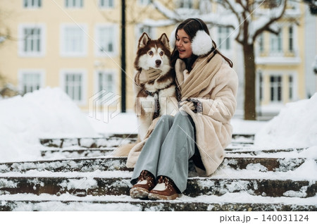 Young woman with long dark hair wearing a beige coat sits on snowy steps with a brown and white husky dog during winter in a city park with buildings in the background Young woman with long dark hair wearing a beige coat sits on snowy steps with a brown and white husky dog during winter in a city park with buildings in the background 140031124