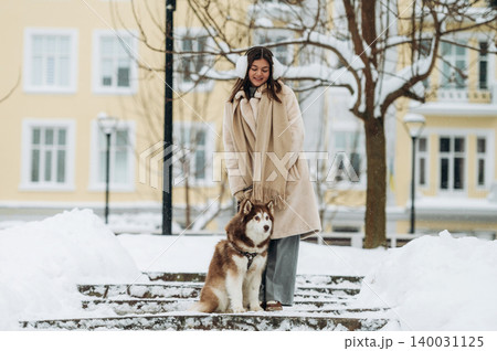 Young woman with long brown hair wearing a beige coat and scarf stands on snowy steps beside a brown and white husky dog in a winter park setting with buildings in the background 140031125