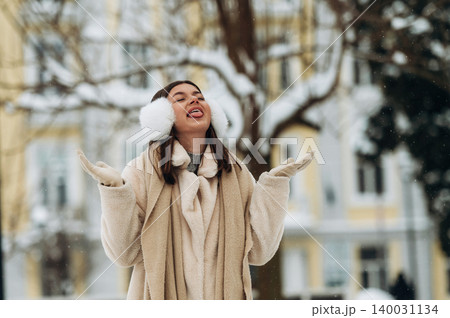 Young woman with brown hair wearing earmuffs and a cozy beige coat is enjoying snowfall outdoors in a winter park with trees and buildings in the background Young woman with brown hair wearing earmuffs and a cozy beige coat is enjoying snowfall outdoors in a winter park with trees and buildings in the background 140031134