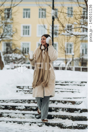 Young woman in a beige coat and scarf is walking down snowy steps while holding a warm drink, with a pastel-colored building in the background during winter 140031136