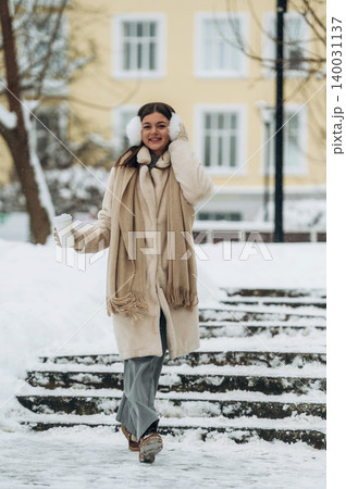 Young woman wearing a cream coat and scarf is walking down snowy steps while holding a warm drink with buildings visible in the background during winter 140031137