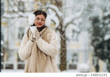 Young woman with brown hair wearing earmuffs and a cozy scarf is holding a warm drink outdoors in a snowy winter setting with blurred trees in the background Young woman with brown hair wearing earmuffs and a cozy scarf is holding a warm drink outdoors in a snowy winter setting with blurred trees in the background 140031141
