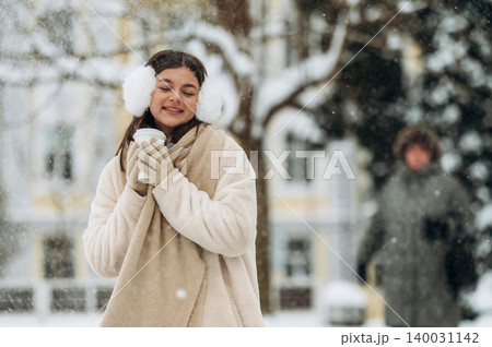 Young woman with earmuffs and gloves is holding a warm drink while enjoying a snowy day in a park with blurred figures and trees in the background during winter season Young woman with earmuffs and gloves is holding a warm drink while enjoying a snowy day in a park with blurred figures and trees in the background during winter season 140031142