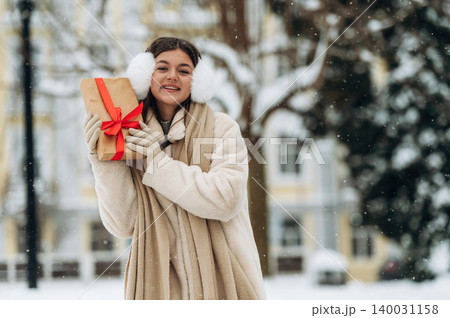 Young woman with brown hair wearing winter clothing is holding a gift box with a red ribbon outdoors in a snowy environment during the holiday season Young woman with brown hair wearing winter clothing is holding a gift box with a red ribbon outdoors in a snowy environment during the holiday season 140031158