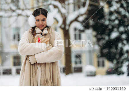 Young woman wearing white earmuffs and a beige coat is holding a gift box while standing in a snowy outdoor setting with trees and buildings in the background Young woman wearing white earmuffs and a beige coat is holding a gift box while standing in a snowy outdoor setting with trees and buildings in the background 140031160