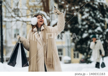 Young woman with long brown hair wearing a cream coat and scarf is taking a selfie while holding shopping bags in a snowy outdoor setting with trees in the background Young woman with long brown hair wearing a cream coat and scarf is taking a selfie while holding shopping bags in a snowy outdoor setting with trees in the background 140031163