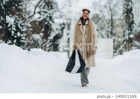 Young woman wearing winter clothing and earmuffs is walking through a snowy path carrying shopping bags in a winter landscape with trees and buildings in the background Young woman wearing winter clothing and earmuffs is walking through a snowy path carrying shopping bags in a winter landscape with trees and buildings in the background 140031169