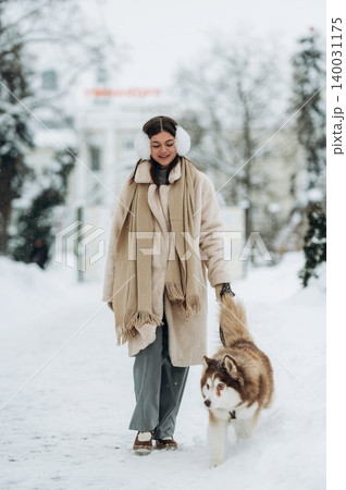 Young woman wearing winter attire and earmuffs walks a fluffy Siberian Husky dog on a snowy path surrounded by trees in a winter landscape during a cold day Young woman wearing winter attire and earmuffs walks a fluffy Siberian Husky dog on a snowy path surrounded by trees in a winter landscape during a cold day 140031175