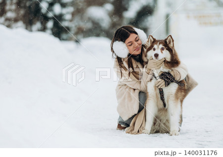 Woman with brown hair wearing earmuffs is hugging a Siberian Husky dog in a snowy outdoor setting, showcasing winter clothing and a serene atmosphere with falling snowflakes Woman with brown hair wearing earmuffs is hugging a Siberian Husky dog in a snowy outdoor setting, showcasing winter clothing and a serene atmosphere with falling snowflakes 140031176