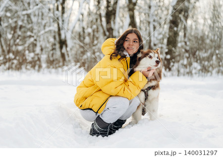 Young girl in a bright yellow winter jacket is crouching in the snow with a fluffy dog, surrounded by a snowy forest landscape during a cold winter day Young girl in a bright yellow winter jacket is crouching in the snow with a fluffy dog, surrounded by a snowy forest landscape during a cold winter day 140031297