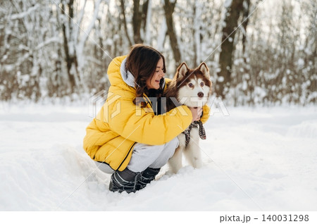 Young woman in a yellow puffer jacket crouches in snow with a Siberian Husky dog, surrounded by a winter forest landscape during a sunny day 140031298