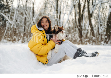 Young girl with long dark hair wearing a yellow winter jacket sits in the snow with a brown and white dog, surrounded by trees in a snowy landscape during winter Young girl with long dark hair wearing a yellow winter jacket sits in the snow with a brown and white dog, surrounded by trees in a snowy landscape during winter 140031308