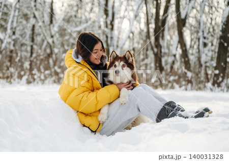 Young woman with dark hair wearing a yellow puffer jacket is sitting in the snow with a brown and white husky dog during a winter day in a snowy forest Young woman with dark hair wearing a yellow puffer jacket is sitting in the snow with a brown and white husky dog during a winter day in a snowy forest 140031328