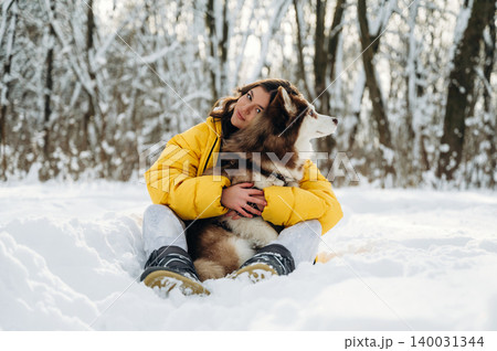 Young woman in a bright yellow jacket sits in the snow with a fluffy brown and white dog, surrounded by a winter forest landscape with snow-covered trees 140031344