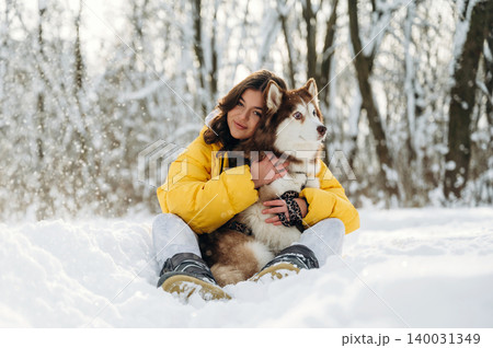Young woman in a bright yellow jacket sits in the snow with a Siberian Husky, surrounded by trees and a winter landscape during a sunny day in a snowy environment Young woman in a bright yellow jacket sits in the snow with a Siberian Husky, surrounded by trees and a winter landscape during a sunny day in a snowy environment 140031349