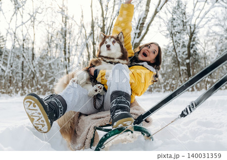 Young woman in yellow jacket joyfully sitting on a sled with a Siberian Husky dog in snowy landscape surrounded by trees during a winter outdoor activity Young woman in yellow jacket joyfully sitting on a sled with a Siberian Husky dog in snowy landscape surrounded by trees during a winter outdoor activity 140031359