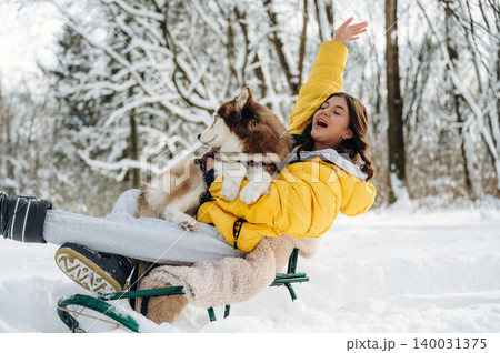 Young woman in a yellow winter jacket is joyfully sitting on a sled with a dog in her arms, surrounded by a snowy landscape and trees during winter fun 140031375