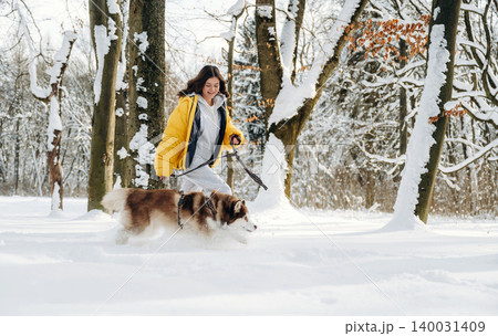 Woman in yellow jacket walking a dog through a snowy forest with trees covered in snow, showcasing a winter outdoor activity in a serene natural environment 140031409
