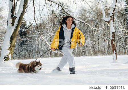 Young woman in a yellow jacket walks a brown and white dog on a snowy path surrounded by trees during winter, showcasing a joyful outdoor activity in a serene landscape Young woman in a yellow jacket walks a brown and white dog on a snowy path surrounded by trees during winter, showcasing a joyful outdoor activity in a serene landscape 140031418