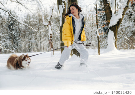 Young woman in a yellow puffer jacket walks a brown and white dog through deep snow in a winter forest, surrounded by trees and a bright, clear sky with copy space Young woman in a yellow puffer jacket walks a brown and white dog through deep snow in a winter forest, surrounded by trees and a bright, clear sky with copy space 140031420