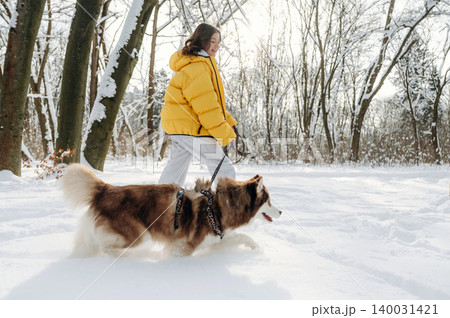 Young woman in a bright yellow puffer jacket walks a fluffy dog on a snowy path surrounded by trees during a winter day in a serene outdoor setting 140031421