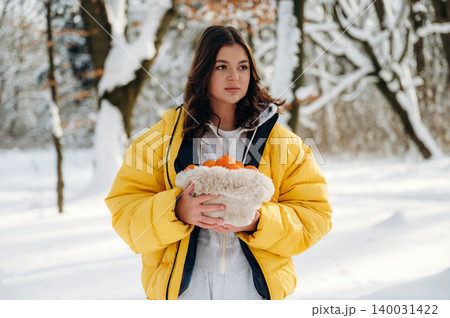 Young woman with long dark hair wearing a yellow puffer jacket is holding a basket of oranges while standing in a snowy forest during winter 140031422