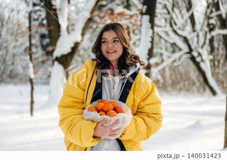 Young woman with curly hair wearing a bright yellow puffer jacket holds a basket of oranges while standing in a snowy forest during winter 140031423