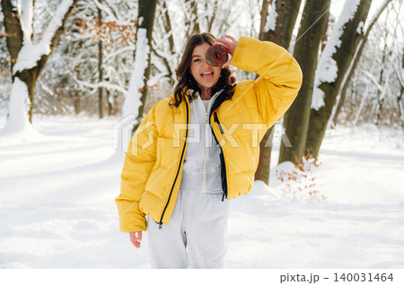 Young woman in a bright yellow puffer jacket is playfully holding a donut while standing in a snowy forest, surrounded by trees and winter scenery with a joyful expression Young woman in a bright yellow puffer jacket is playfully holding a donut while standing in a snowy forest, surrounded by trees and winter scenery with a joyful expression 140031464