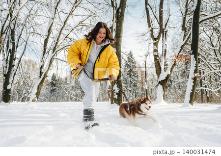 Young woman in a yellow puffer jacket is walking a brown and white dog through a snowy park with trees in the background during wintertime outdoor activity Young woman in a yellow puffer jacket is walking a brown and white dog through a snowy park with trees in the background during wintertime outdoor activity 140031474