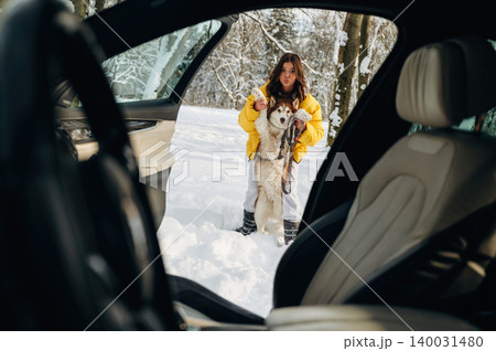 Young woman in a yellow winter jacket stands outside a car holding a dog in the snow, surrounded by trees in a winter landscape with a cozy atmosphere Young woman in a yellow winter jacket stands outside a car holding a dog in the snow, surrounded by trees in a winter landscape with a cozy atmosphere 140031480
