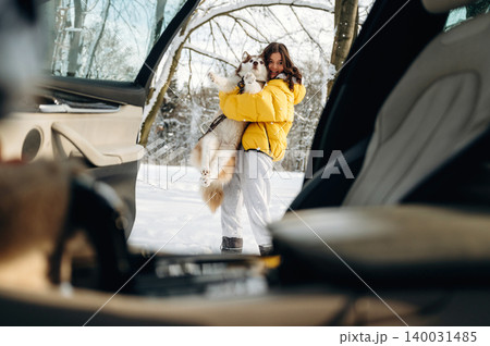 Young girl in a yellow jacket is holding a fluffy dog while standing outside a car in a snowy landscape with trees in the background during winter 140031485