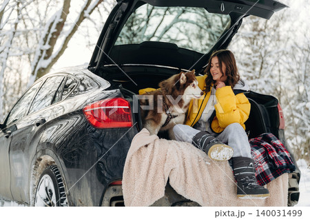 Young woman in a yellow jacket sitting in the trunk of a black SUV with a brown and white dog during winter in a snowy forest setting with trees in the background Young woman in a yellow jacket sitting in the trunk of a black SUV with a brown and white dog during winter in a snowy forest setting with trees in the background 140031499