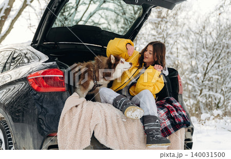Young woman in yellow jacket sitting in car trunk with her dog during winter, snow-covered landscape and trees visible in the background, cozy atmosphere with blanket Young woman in yellow jacket sitting in car trunk with her dog during winter, snow-covered landscape and trees visible in the background, cozy atmosphere with blanket 140031500