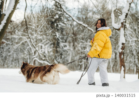 Young woman wearing a bright yellow puffer jacket walks her fluffy dog on a snowy path surrounded by trees, showcasing a winter outdoor scene with a playful atmosphere Young woman wearing a bright yellow puffer jacket walks her fluffy dog on a snowy path surrounded by trees, showcasing a winter outdoor scene with a playful atmosphere 140031515