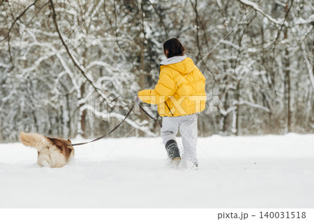 Child wearing a bright yellow puffer jacket walks a dog on a snowy path surrounded by trees in a winter landscape with soft snow covering the ground Child wearing a bright yellow puffer jacket walks a dog on a snowy path surrounded by trees in a winter landscape with soft snow covering the ground 140031518