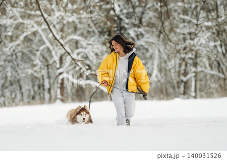 Young woman in a yellow puffer jacket walks a dog on a leash through a snowy landscape with trees in the background during winter outdoor activity 140031526