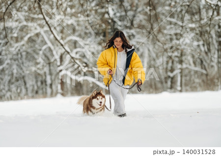Young woman in a bright yellow puffer jacket walks her dog through a snowy landscape, surrounded by trees and a winter wonderland atmosphere during a leisurely stroll Young woman in a bright yellow puffer jacket walks her dog through a snowy landscape, surrounded by trees and a winter wonderland atmosphere during a leisurely stroll 140031528