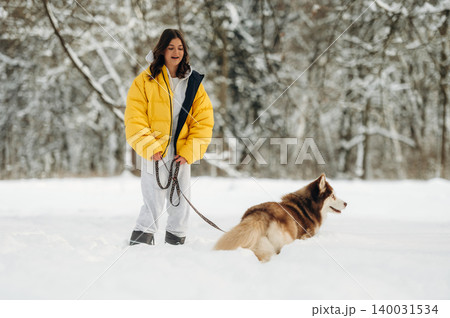Young woman in a bright yellow puffer jacket walks a brown and white husky dog on a snowy path surrounded by trees in a winter landscape with soft sunlight Young woman in a bright yellow puffer jacket walks a brown and white husky dog on a snowy path surrounded by trees in a winter landscape with soft sunlight 140031534
