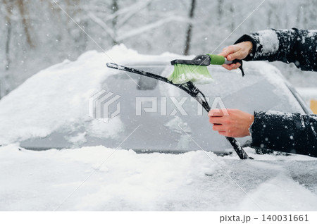 Person using a snow brush to clear snow from a car windshield during a winter snowfall, with a blurred snowy landscape in the background and falling snowflakes visible 140031661