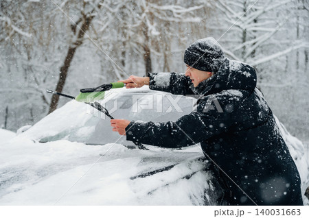 Man in black winter coat is clearing snow from a car windshield using a green snow brush during a snowy day in a winter landscape with trees in the background 140031663