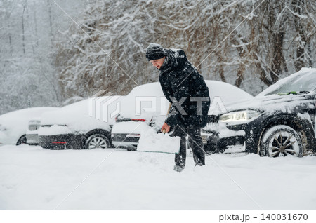 Man in black coat and hat shoveling snow in a parking lot covered with snow, several cars partially buried in the background during winter weather conditions 140031670