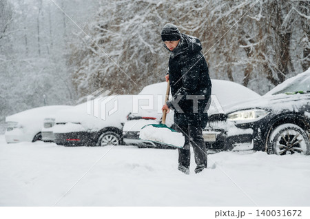 Individual in black winter coat shoveling snow in a parking lot covered in snow with parked cars partially obscured by snow during a winter storm 140031672