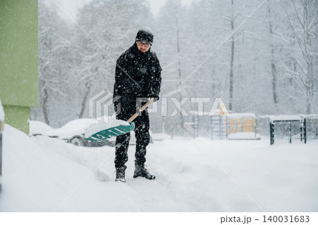Man in black winter clothing shoveling snow with a green shovel during a heavy snowfall near a playground and parked cars in a residential area Man in black winter clothing shoveling snow with a green shovel during a heavy snowfall near a playground and parked cars in a residential area 140031683