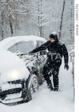 Man dressed in black winter clothing is clearing snow from a car parked in a snowy landscape with trees and heavy snowfall creating a winter scene 140031702